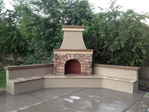 Outdoor stone fireplace with built-in benches, surrounded by greenery, on a concrete patio.