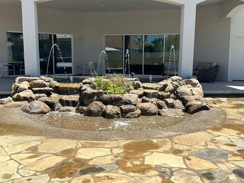 Water fountain with stacked rocks and jets on a stone patio in front of a building with glass doors.