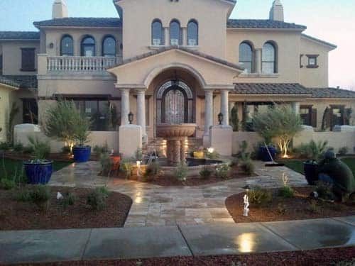 Two-story beige house with fountain, pathway, and landscaping; a person is gardening.
