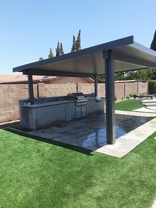 Outdoor kitchen under a gray sloped awning. Grill, counter, and cabinets on a concrete patio, bordered by artificial turf.