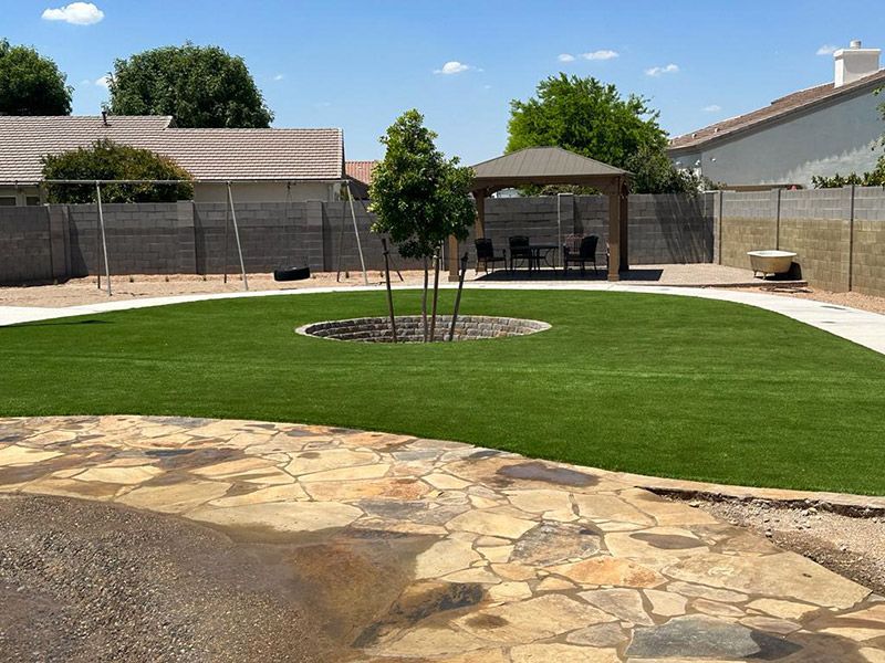Lush green backyard with stone patio, artificial turf, gazebo, small trees, and a swing set. Sunny day.
