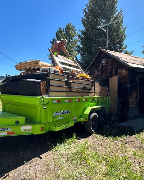 A green trailer filled with junk is parked in front of a shed.