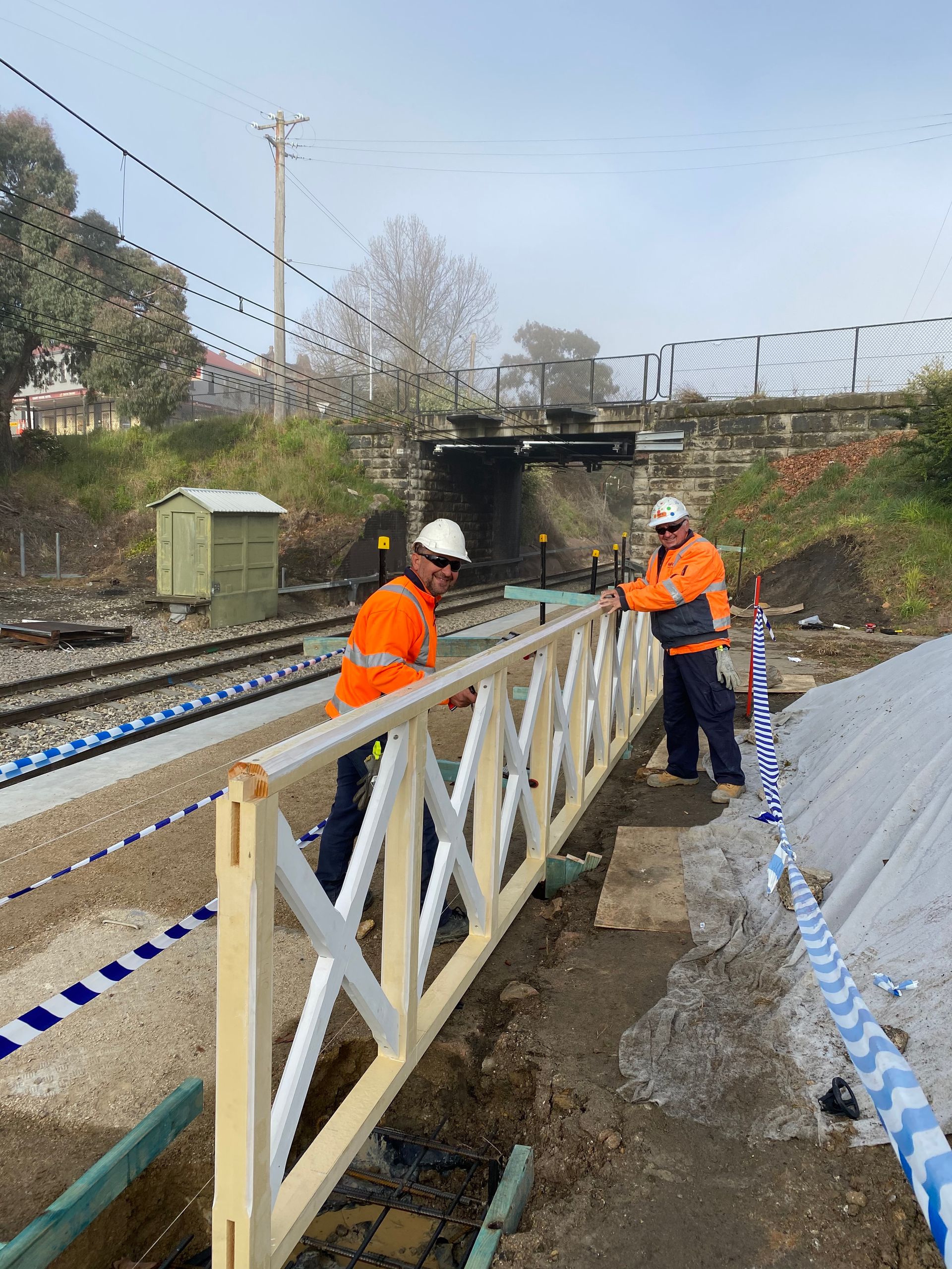 Two construction workers are working on a fence near a train track.