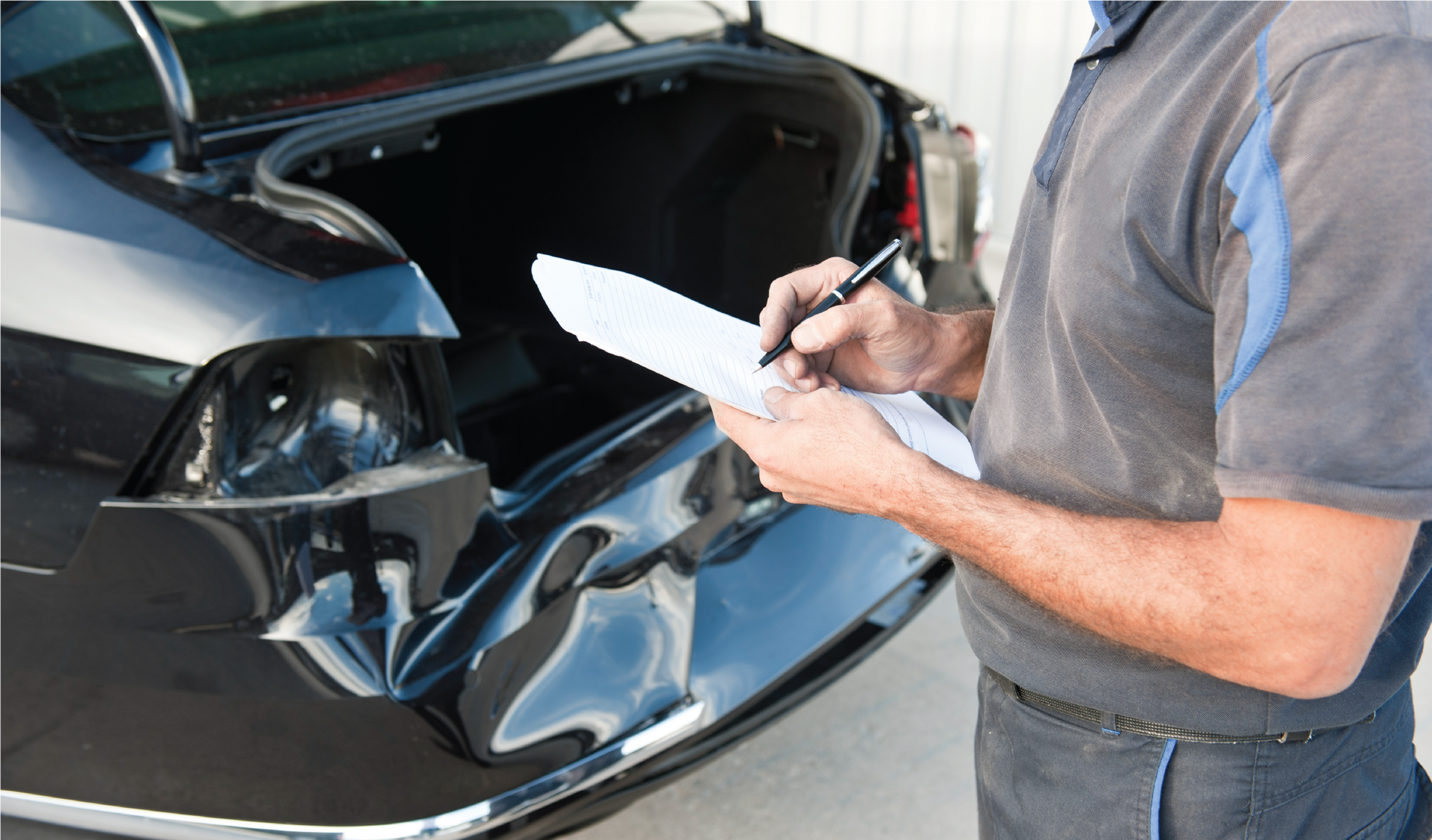 A man is writing on a clipboard in front of a damaged car.