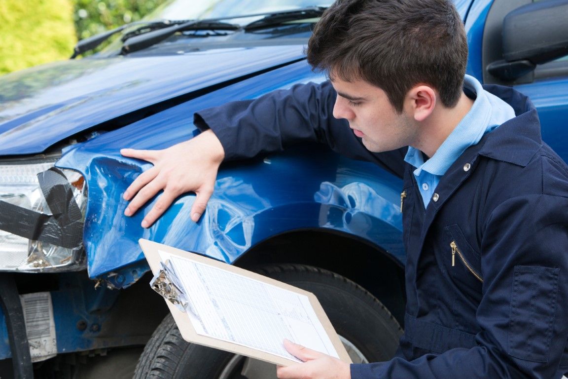 Man inspecting front damage on a blue car after a crash, holding a clipboard