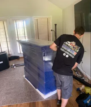 A man in a black shirt is wrapping a dresser in blue plastic wrap.