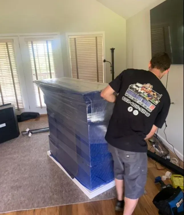 Man wraps a large, blue-wrapped item with plastic wrap inside a room with wood floors and windows.