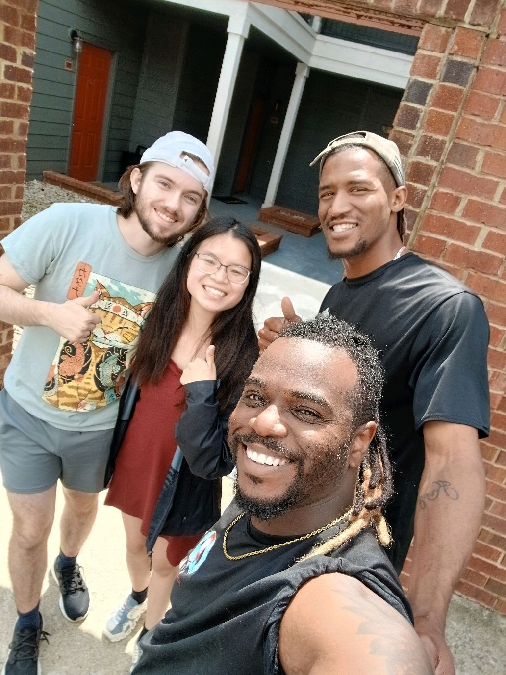 Four people smiling, posing for a selfie outdoors, near a brick wall.