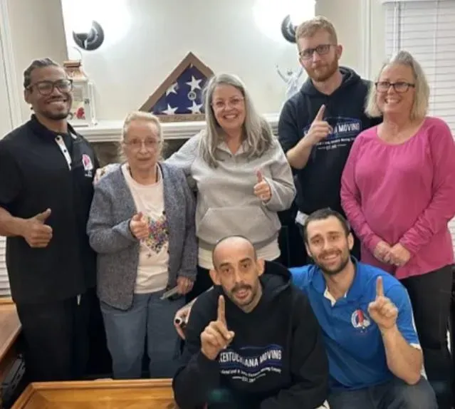 Group of people smiling and giving thumbs up. Living room setting, flag in background.