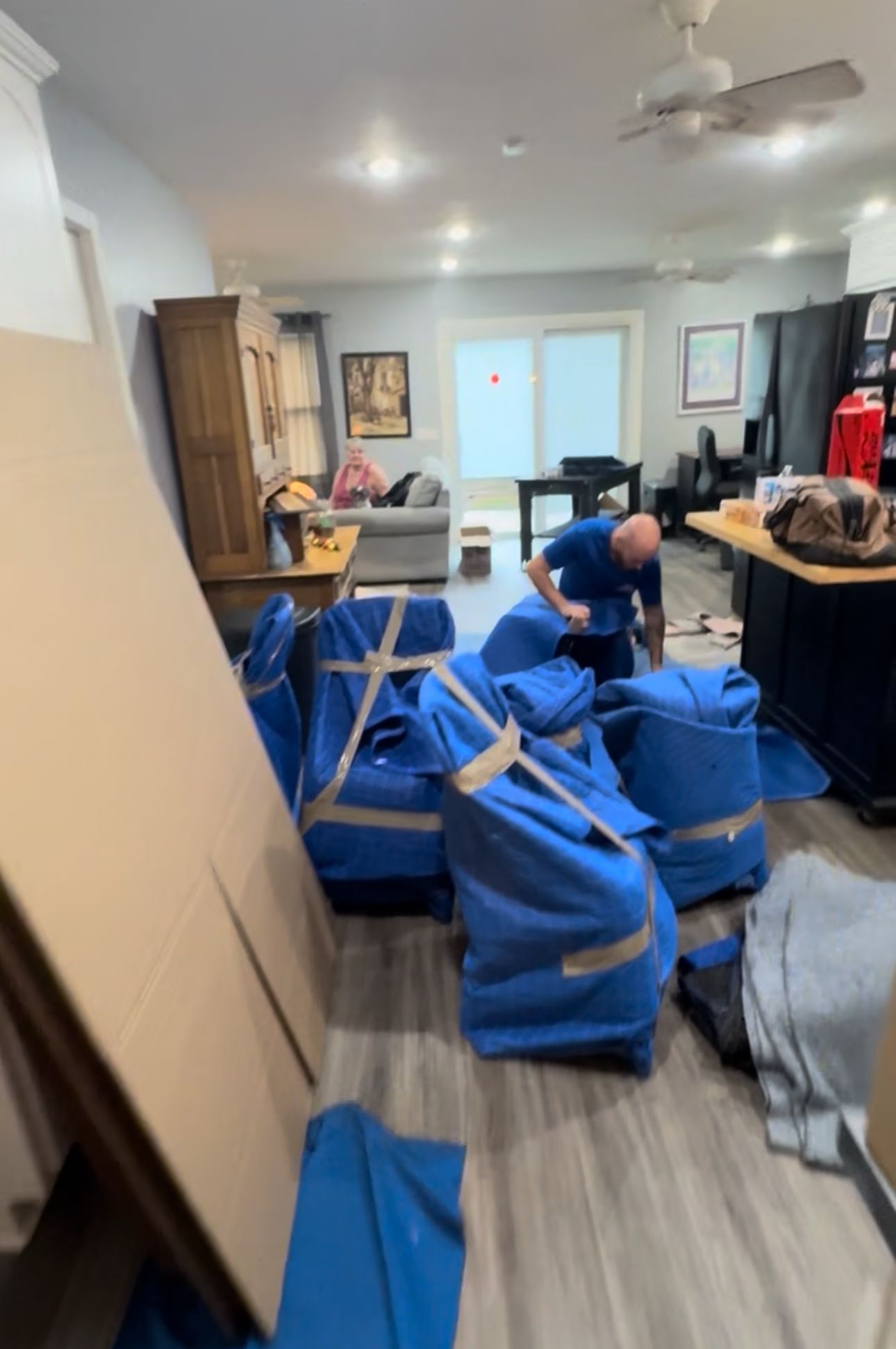 A man packages items inside a home during a move. Blue bags and cardboard surround him; other furniture, and a couch are visible.