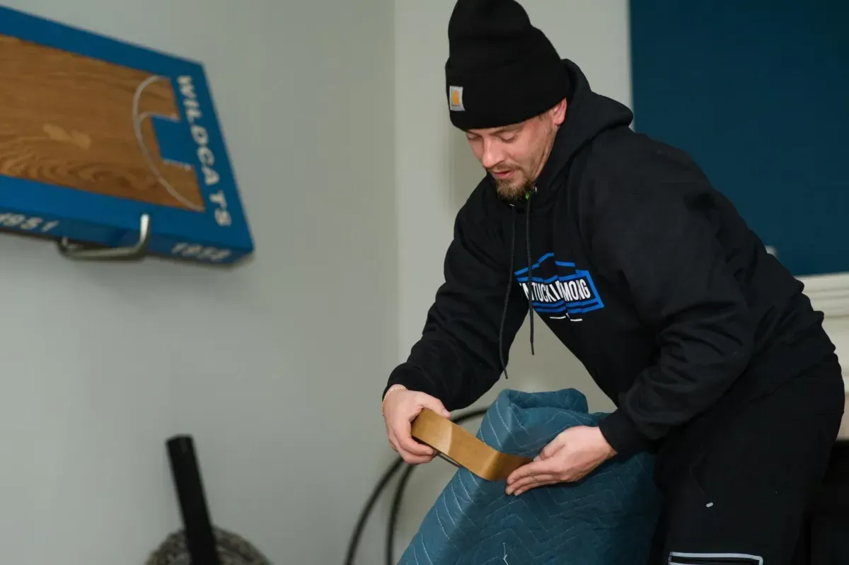 Man taping a piece of furniture, wearing a black beanie and hoodie, inside a room with a blue basketball hoop.