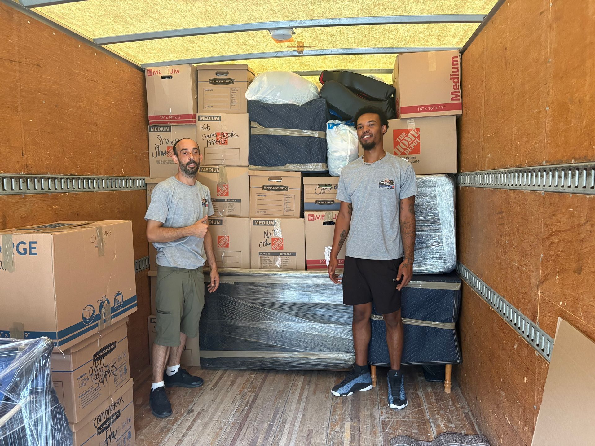 Two movers stand inside a moving truck filled with boxes and furniture. One gives a thumbs up.