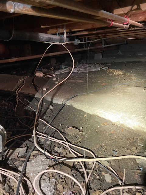 Under-house crawl space with electrical wires, concrete and dirt floor, wood beams and pipes.