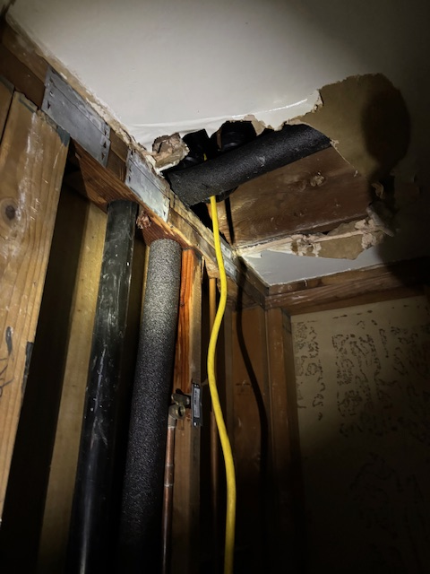 Damaged ceiling exposing pipes, wiring, and wooden beams. Interior shot with a yellow wire hanging.