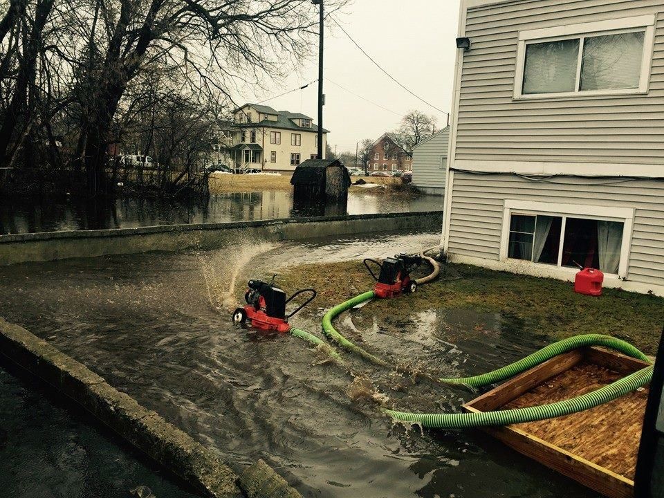 Two red pumps spraying water onto a flooded street, beside a gray building. Green hoses and a wooden box are visible.