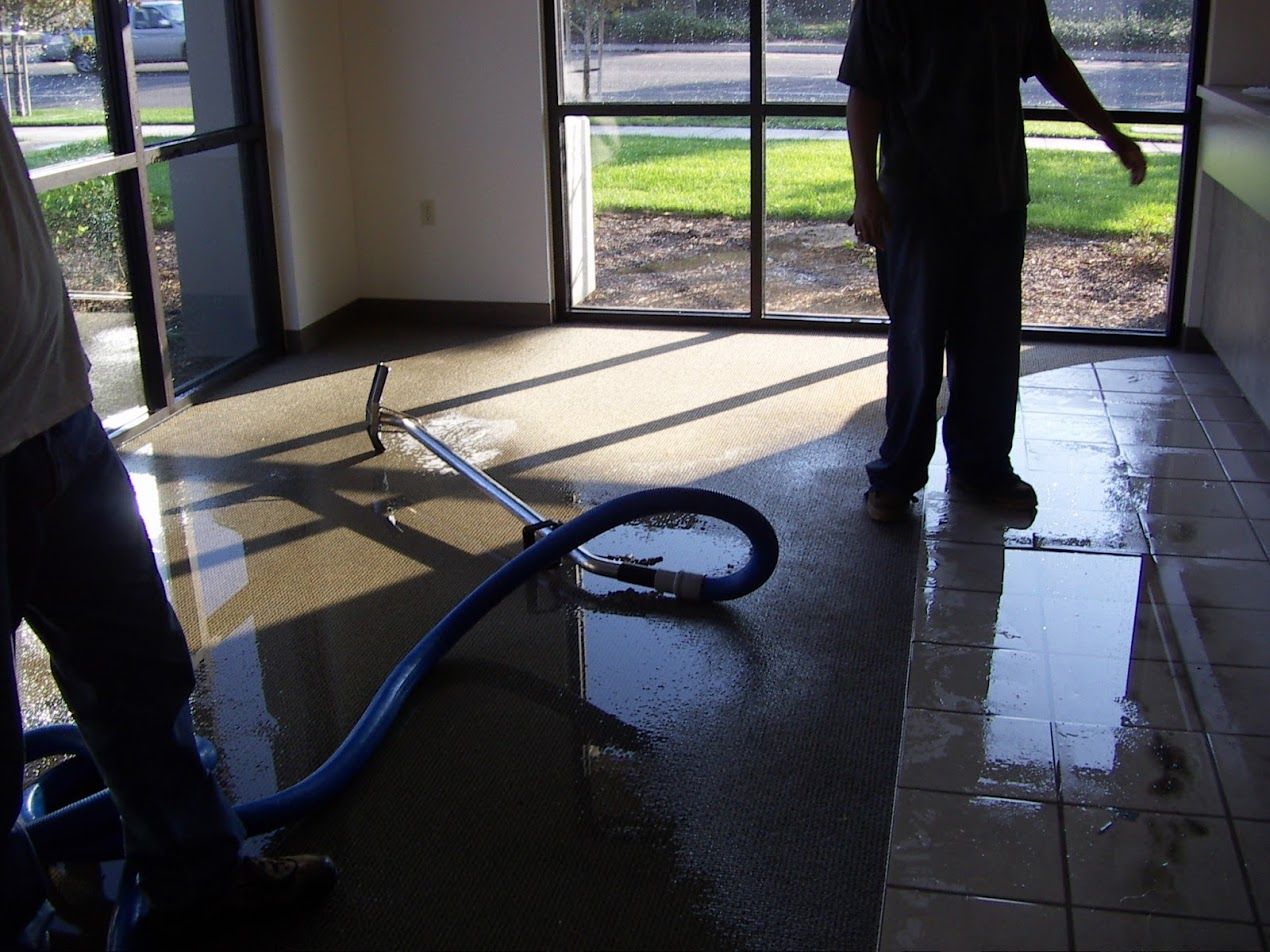 Workers cleaning a flooded commercial space with a hose. Water covers floor; sunlight streams through windows.