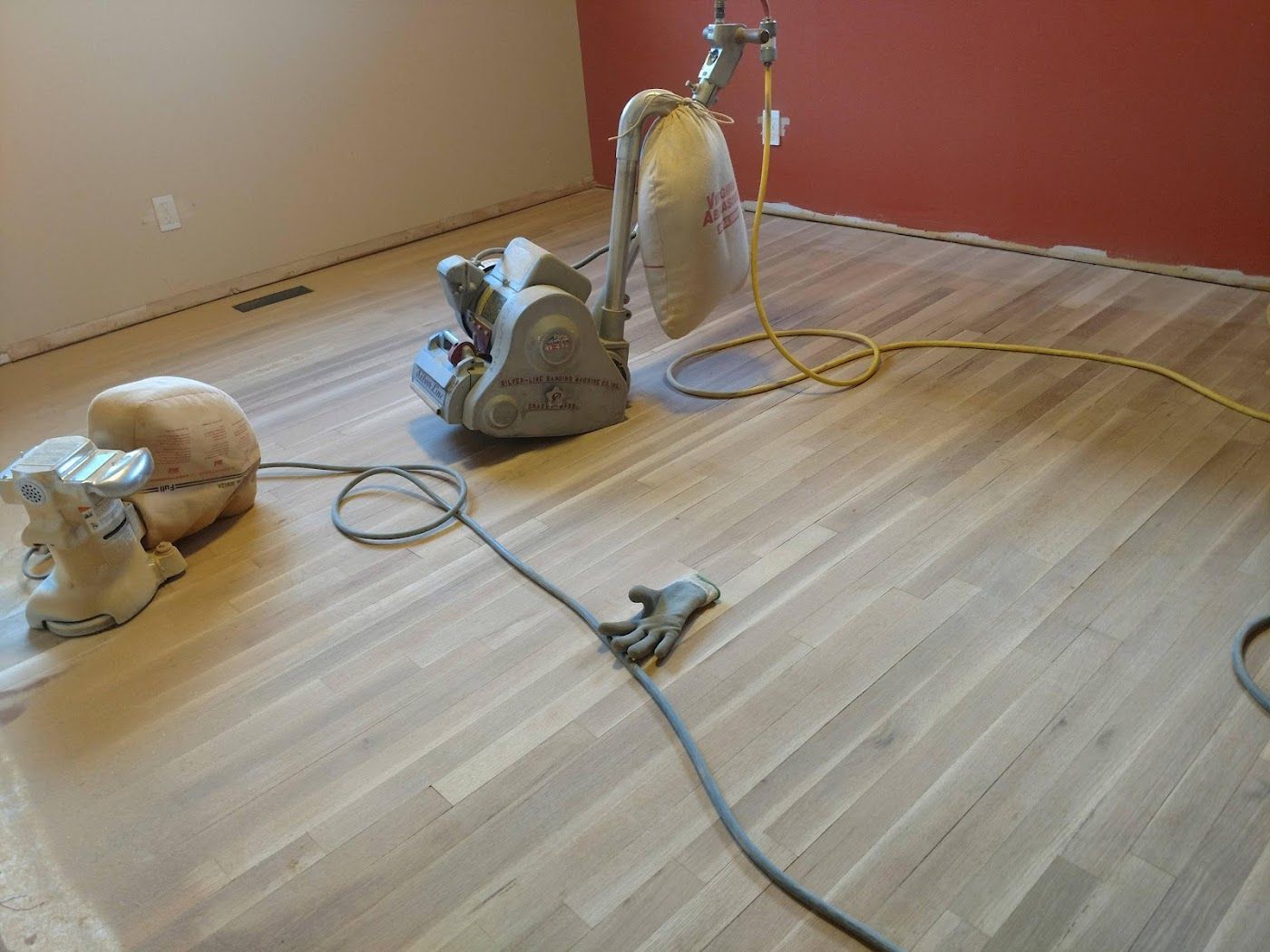 Floor sanders on hardwood floor, beige and red walls in the background. Sanding is in progress.