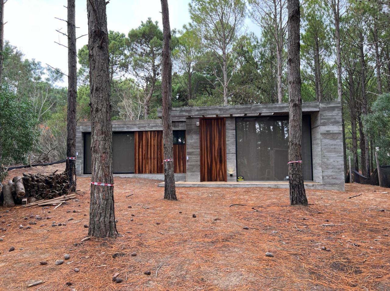 Moderna estructura de hormigón con puertas color óxido, ubicada en un bosque con pinos y vegetación baja marrón.