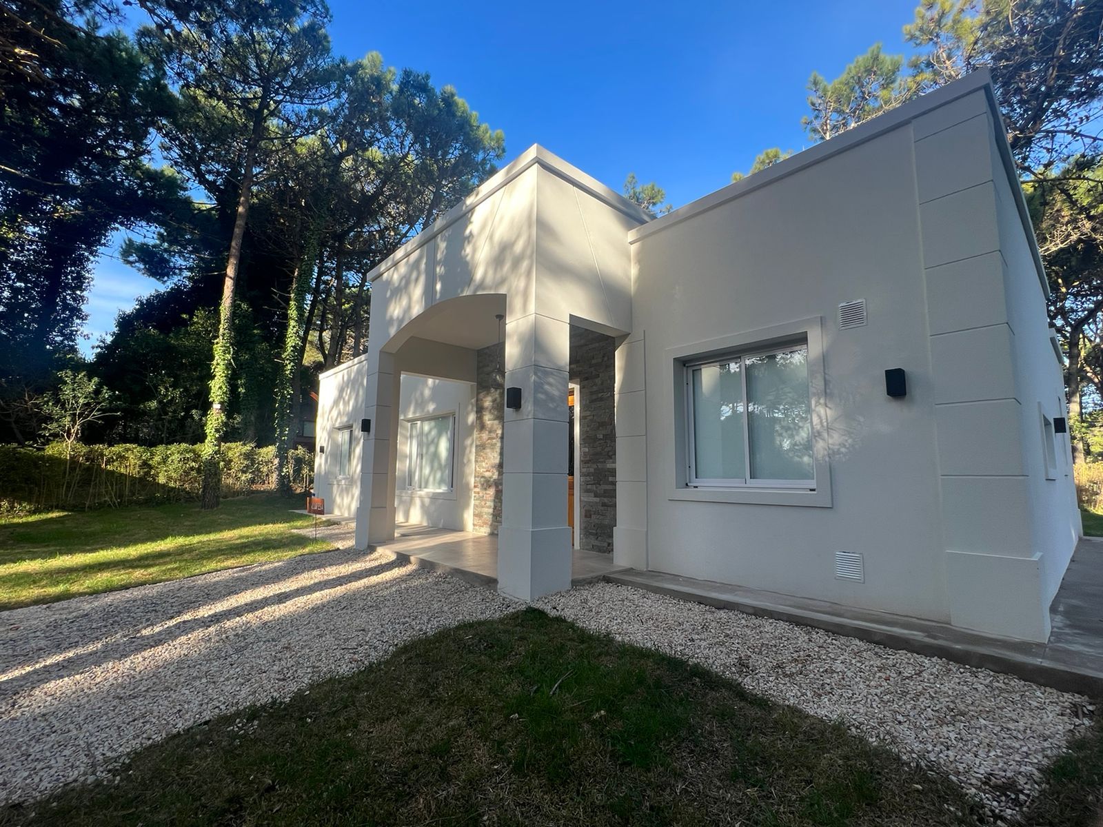 Casa moderna blanca con entrada de grava y árboles alrededor.