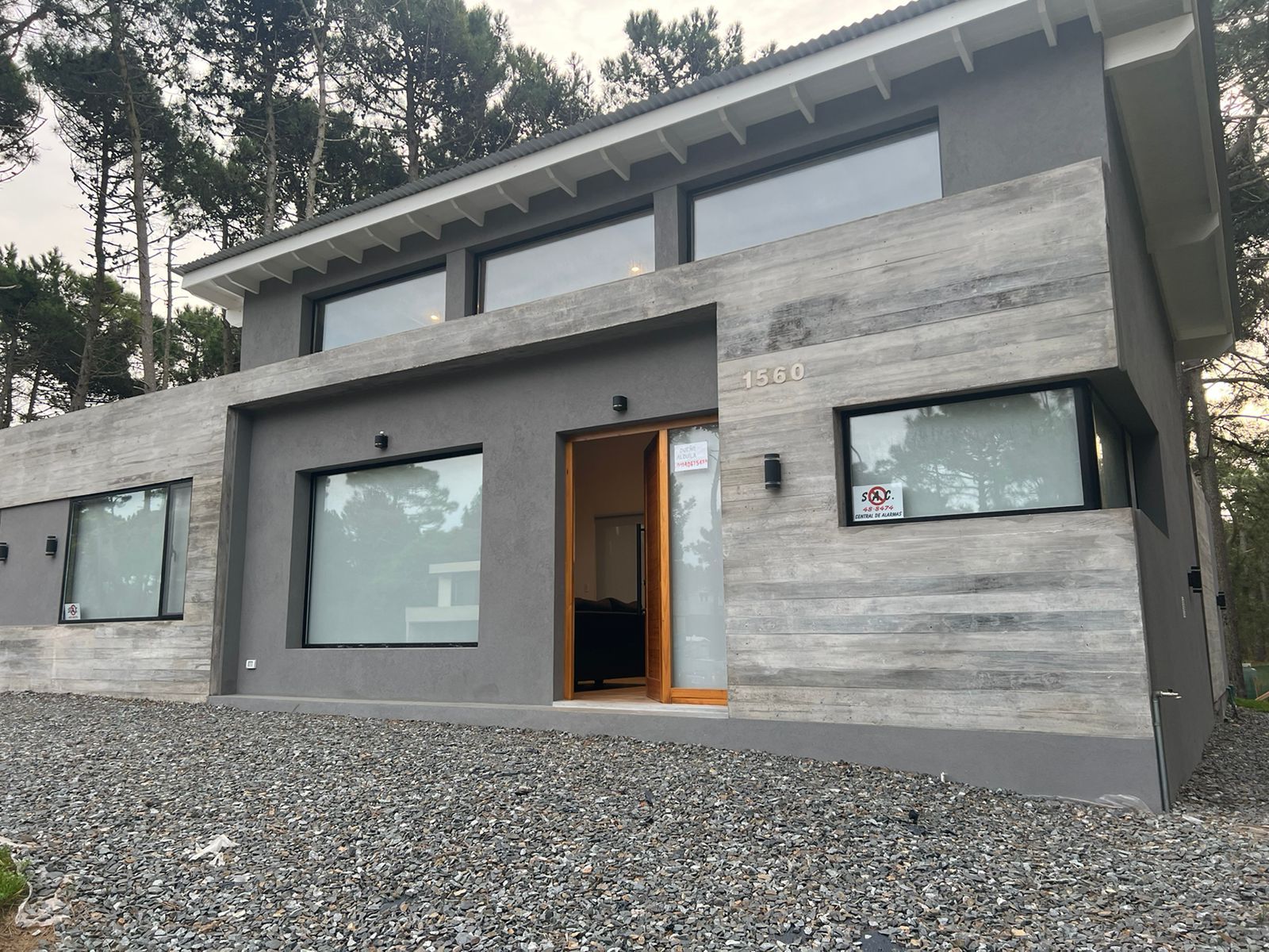 Moderna casa de dos plantas con fachada de hormigón gris y grandes ventanales, rodeada de árboles y con camino de grava.