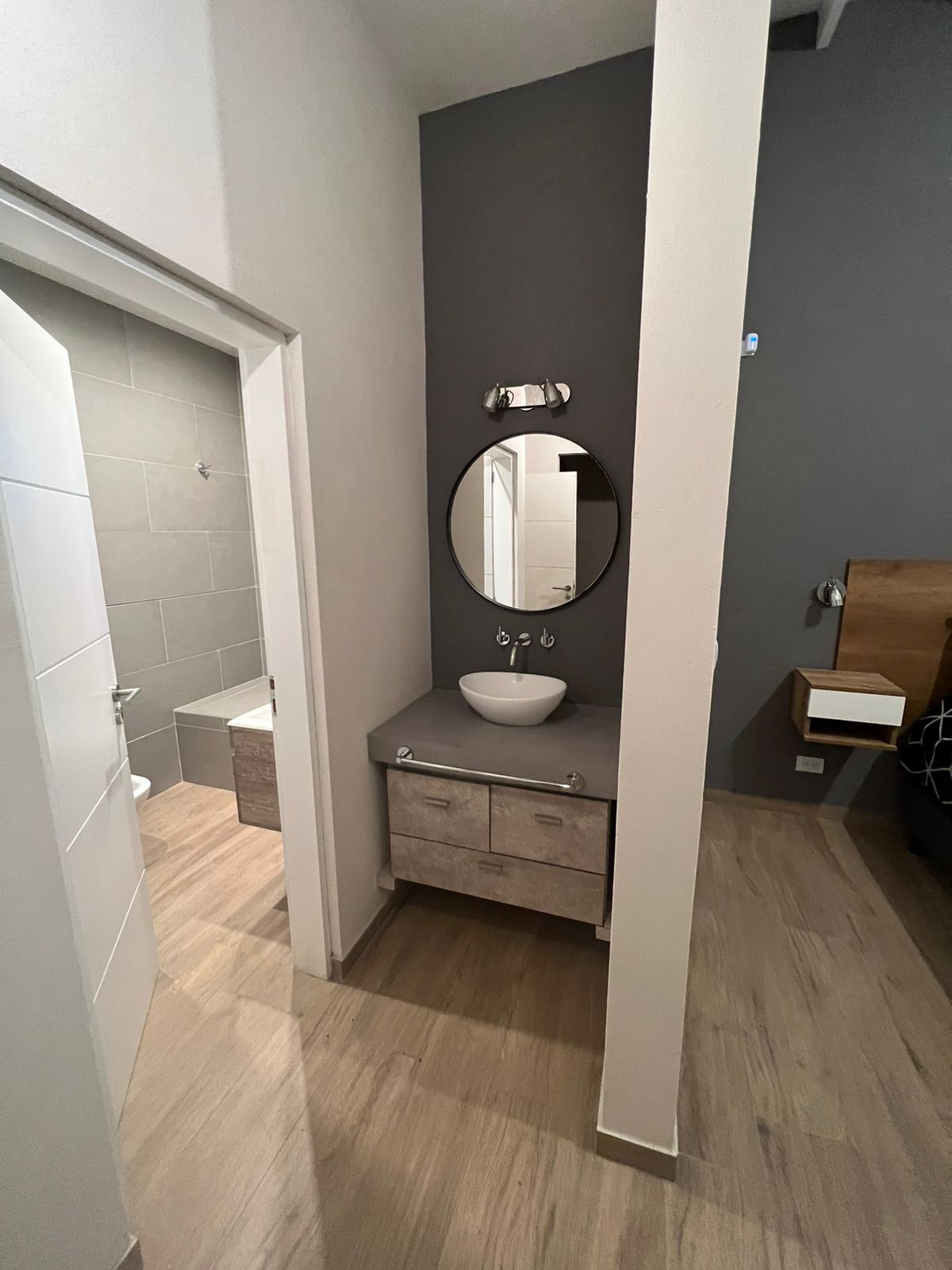 Baño con tocador, espejo redondo y puerta al área del inodoro; colores neutros y suelos de madera clara.