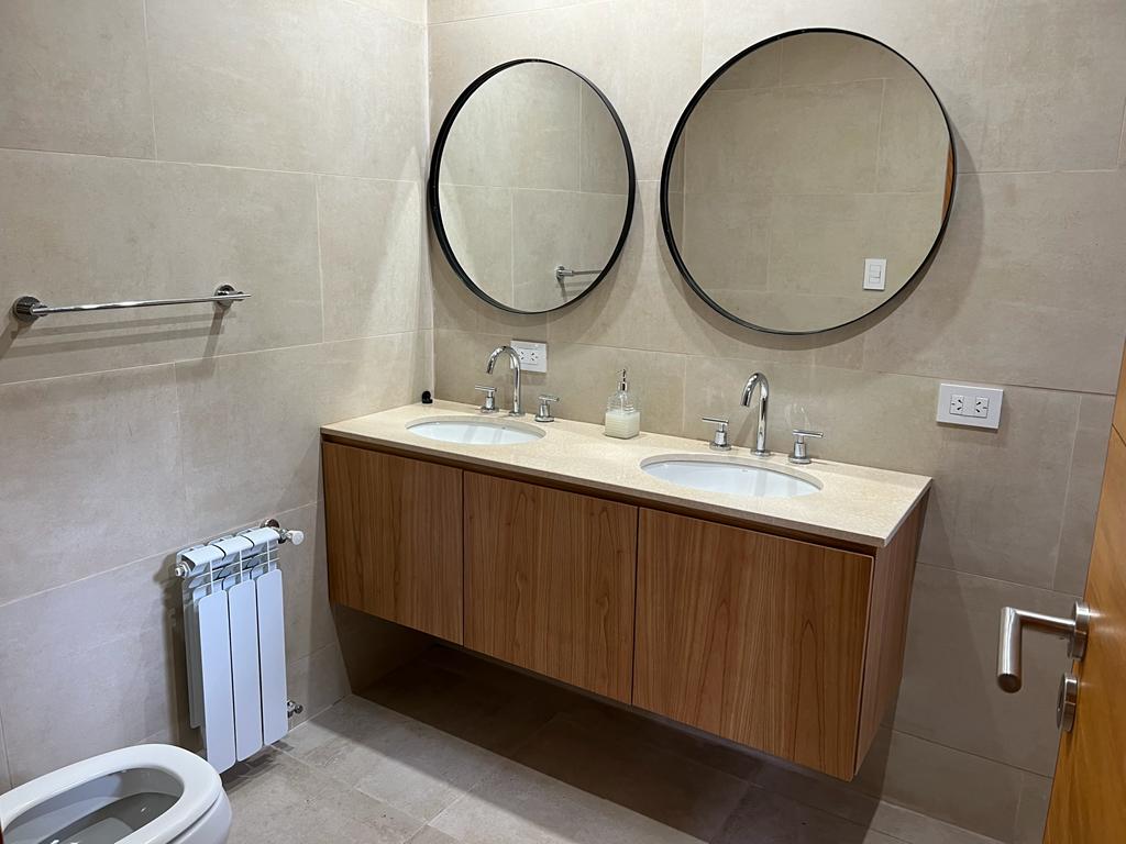 Baño con lavabo doble, espejos redondos, tocador de madera y azulejos color beige.