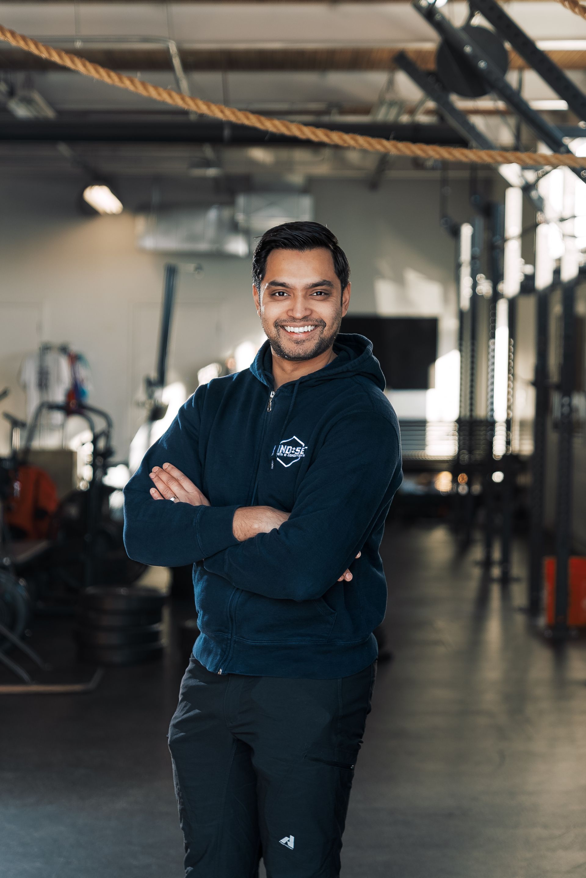 A man is standing in a gym with his arms crossed and smiling.