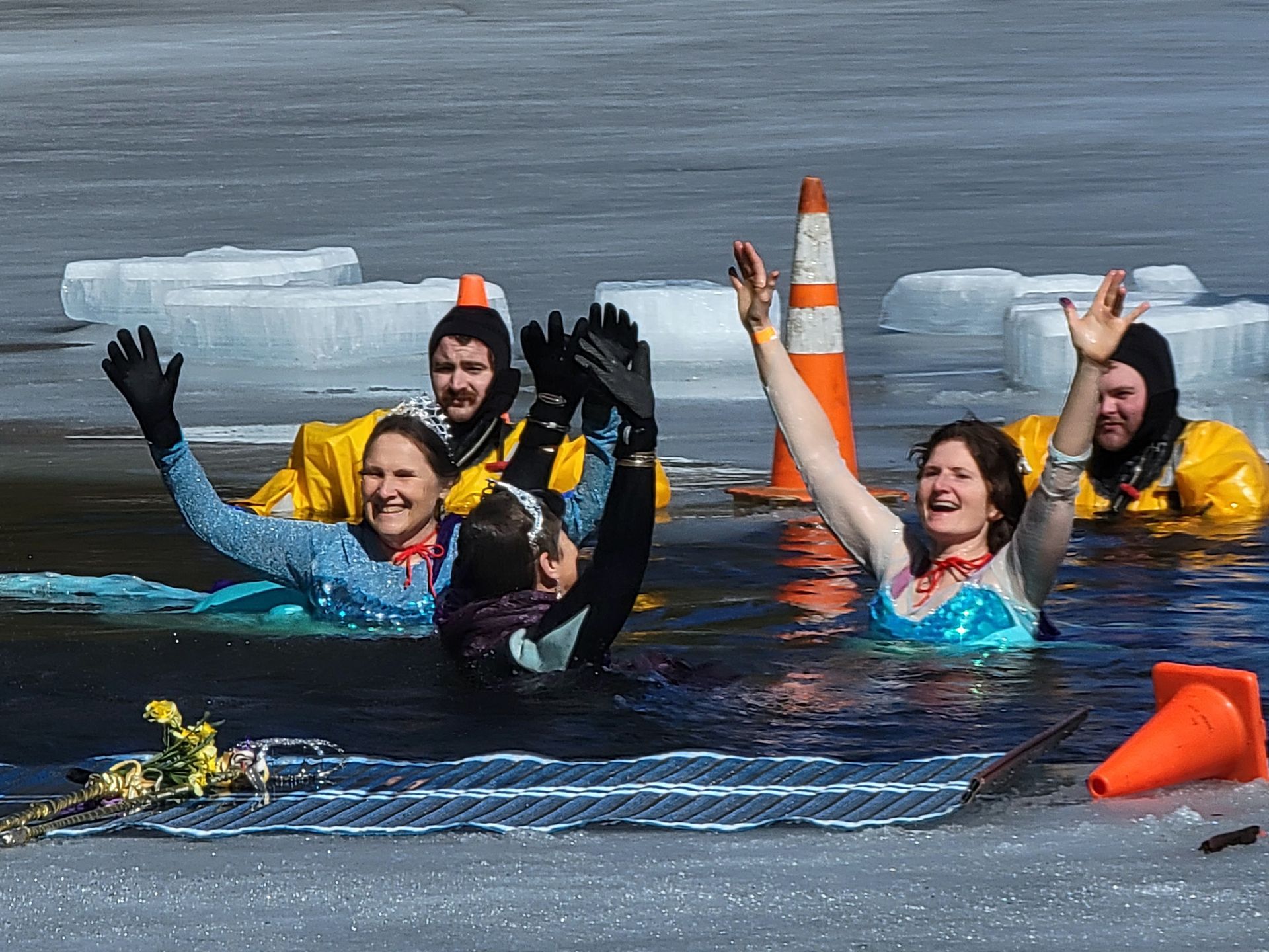 Image shows 4 women, dressed as princesses, with their hands up in the air and smiling in water surrounded by ice.  2 firefighters in cold water rescue suits look on from the water.