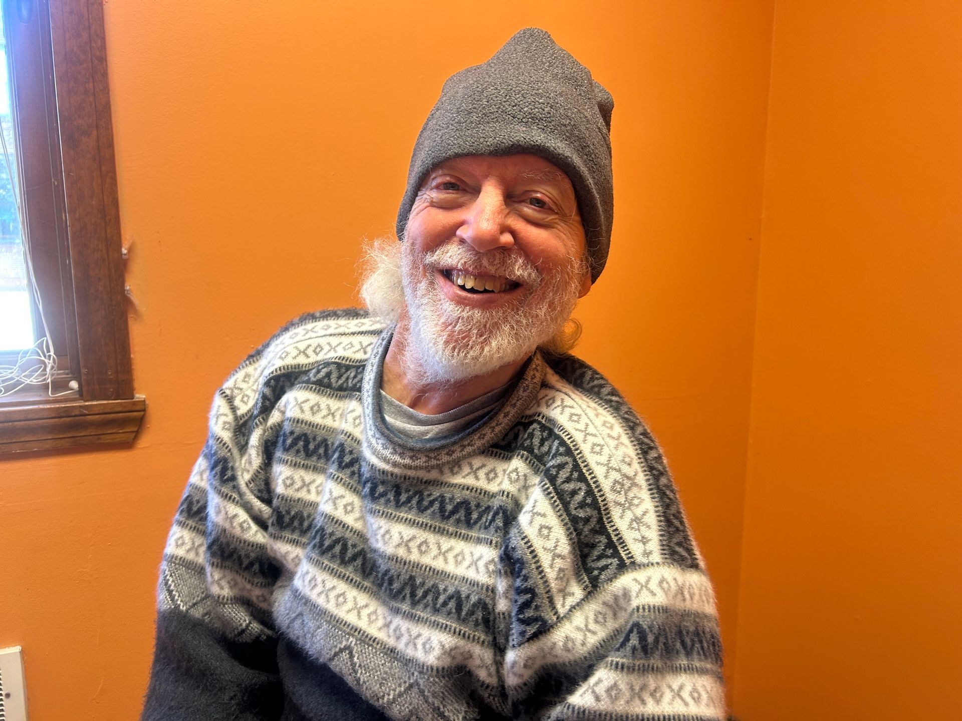 Image is of Brian Adams, wearing a grey winter hat and smiling broadly with an orange background