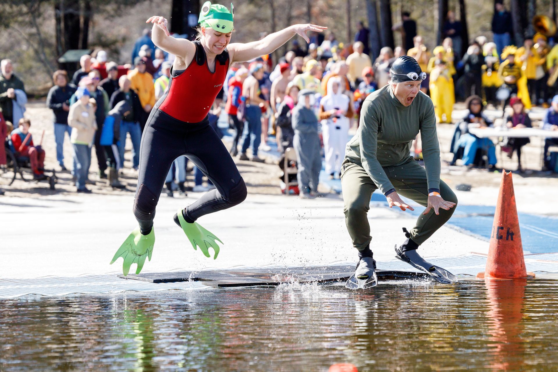 Image shows 2 women, dressed as frogs, frog-hopping into water surrounded by ice.  A large crowd is in the background.