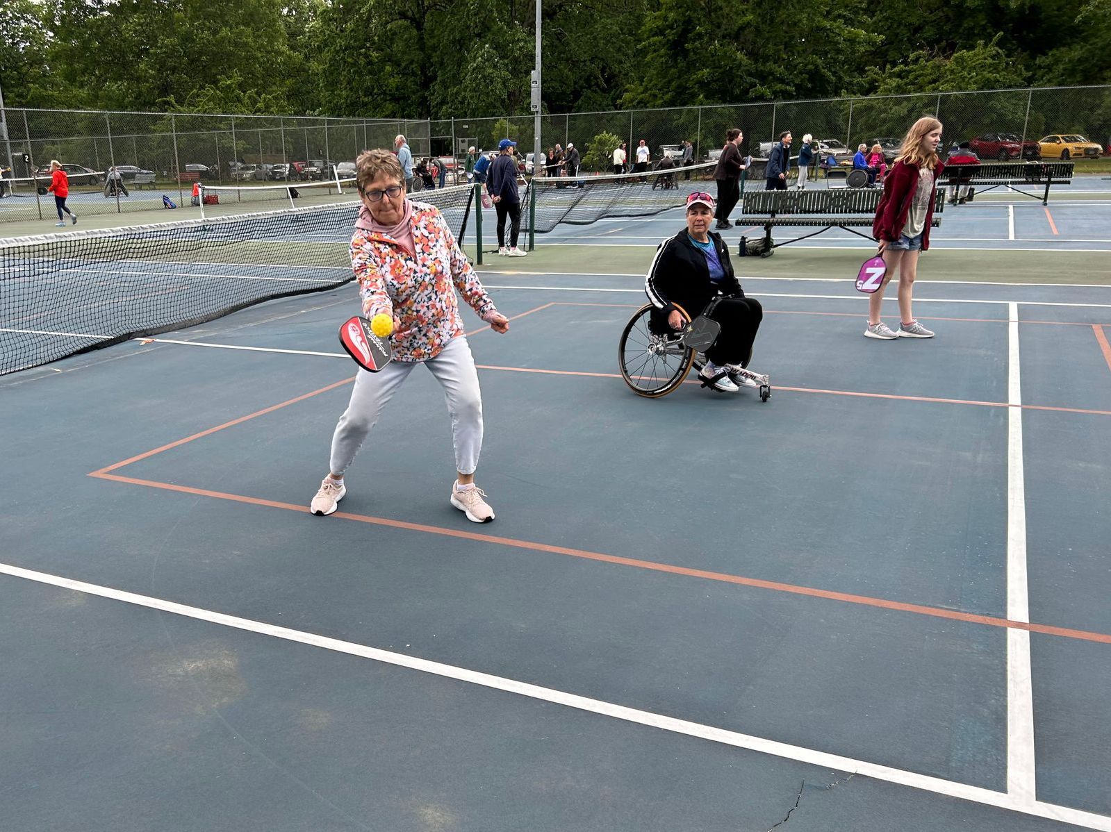 Image shows people playing pickleball; a person standing is about to hit the ball while a wheelchair user looks on, ready