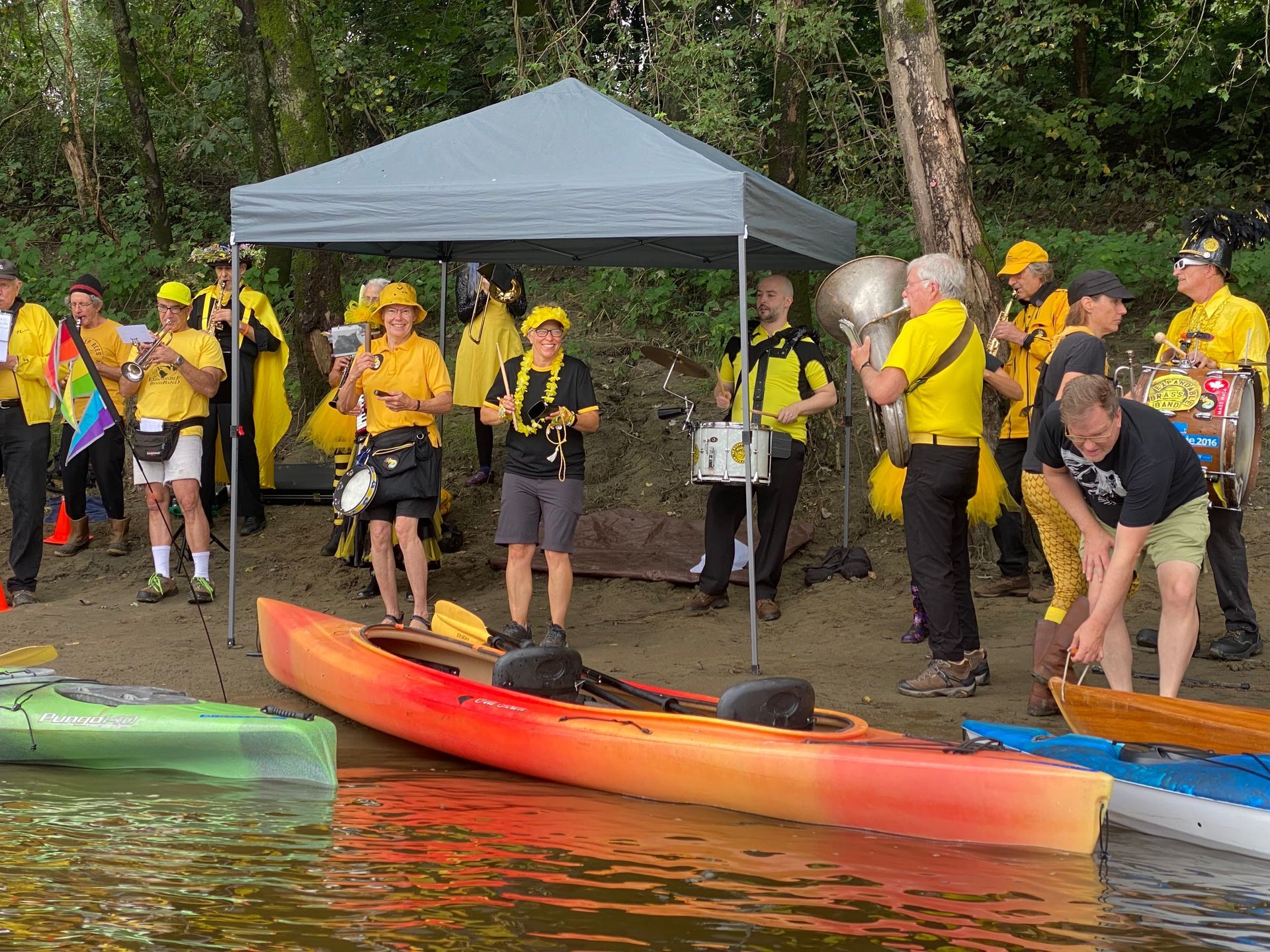 Image shows a band dressed in yellow and black, playing on the riverbank; kayaks are in front of them