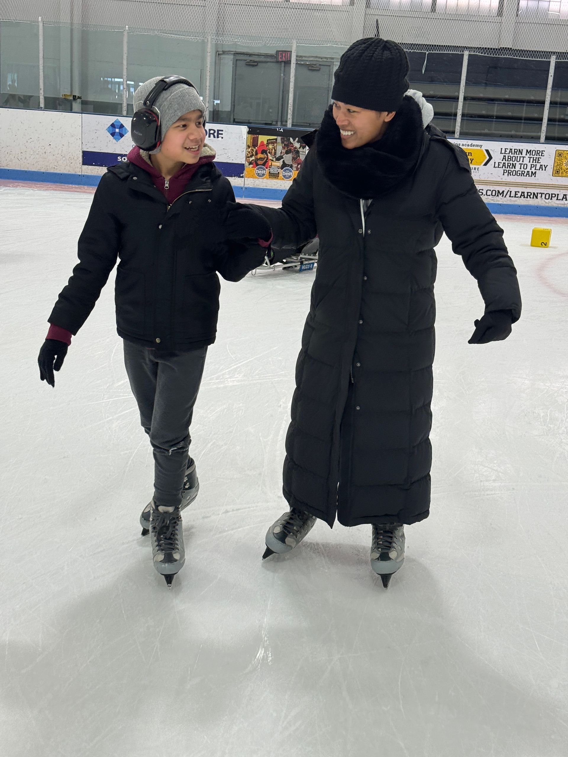 2 people are stand-skating, smiling at each other; one is wearing noise-canceling headphones