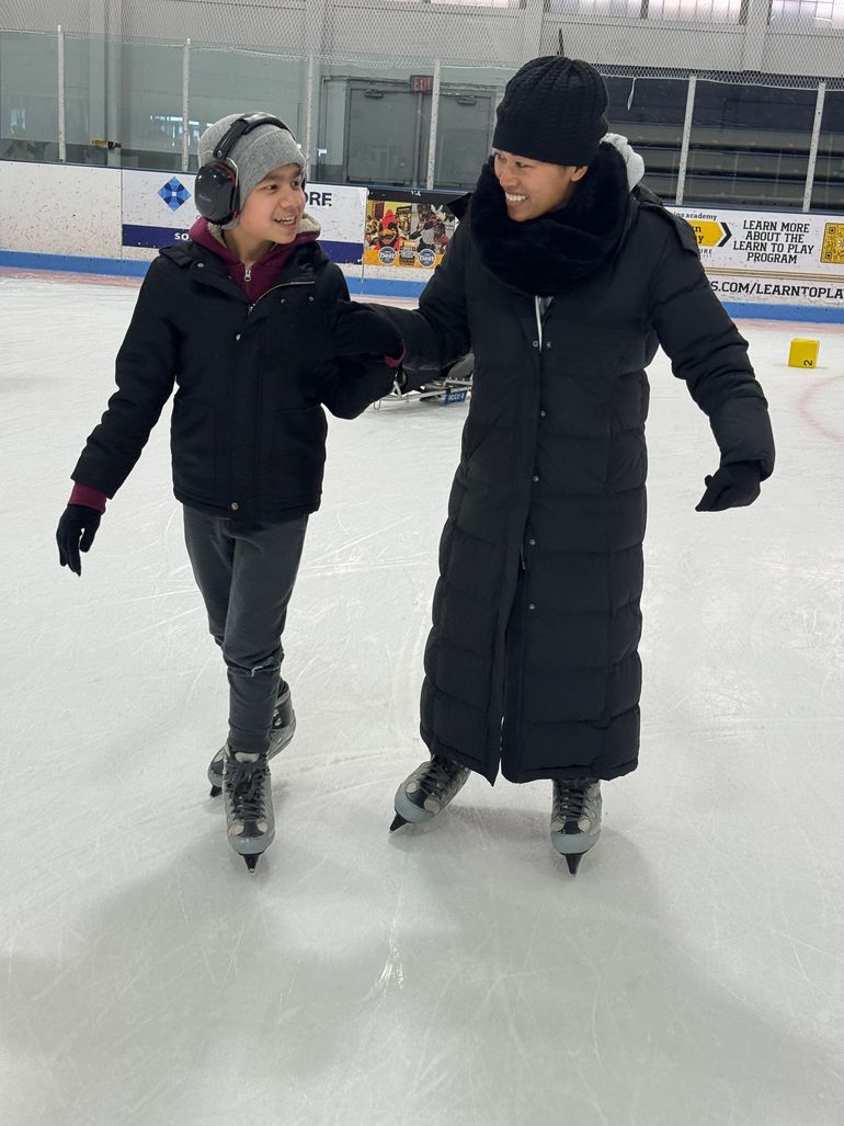 A teenager and a woman, hold hands while ice skating.  They are looking at each other and smiling; the teenager is wearing noise canceling headphones.