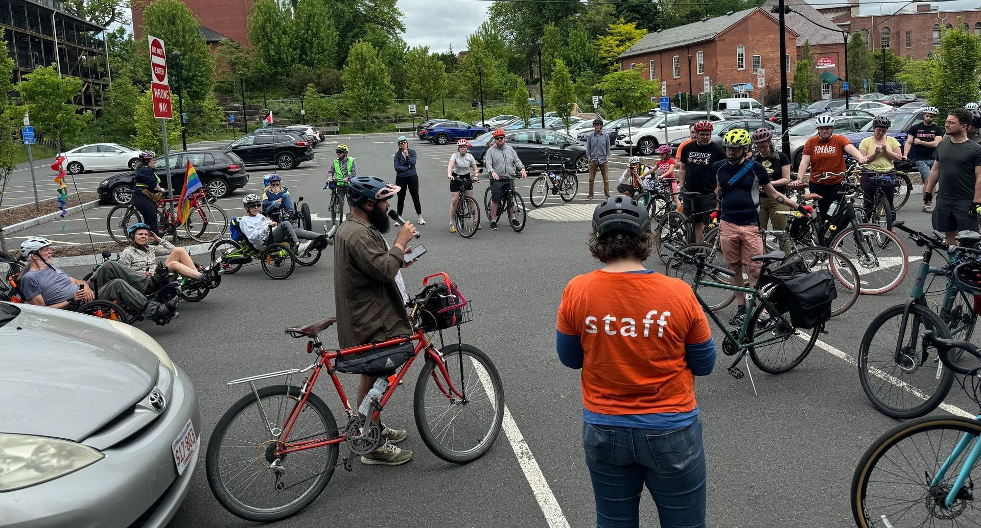 An All Out Adventures program leader holds a clipboard while another leader gives instructions to a large group of cyclists