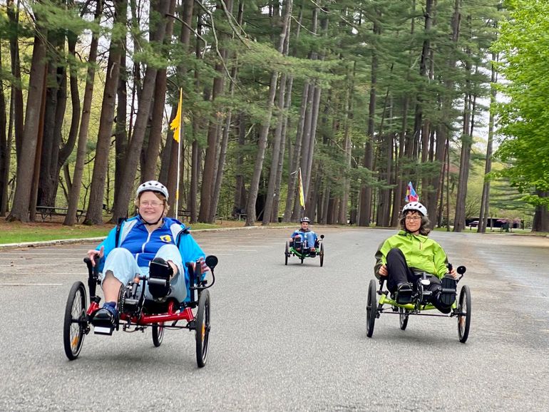 A group of 3 recumbent cyclists pedal together on an empty road; all are smiling