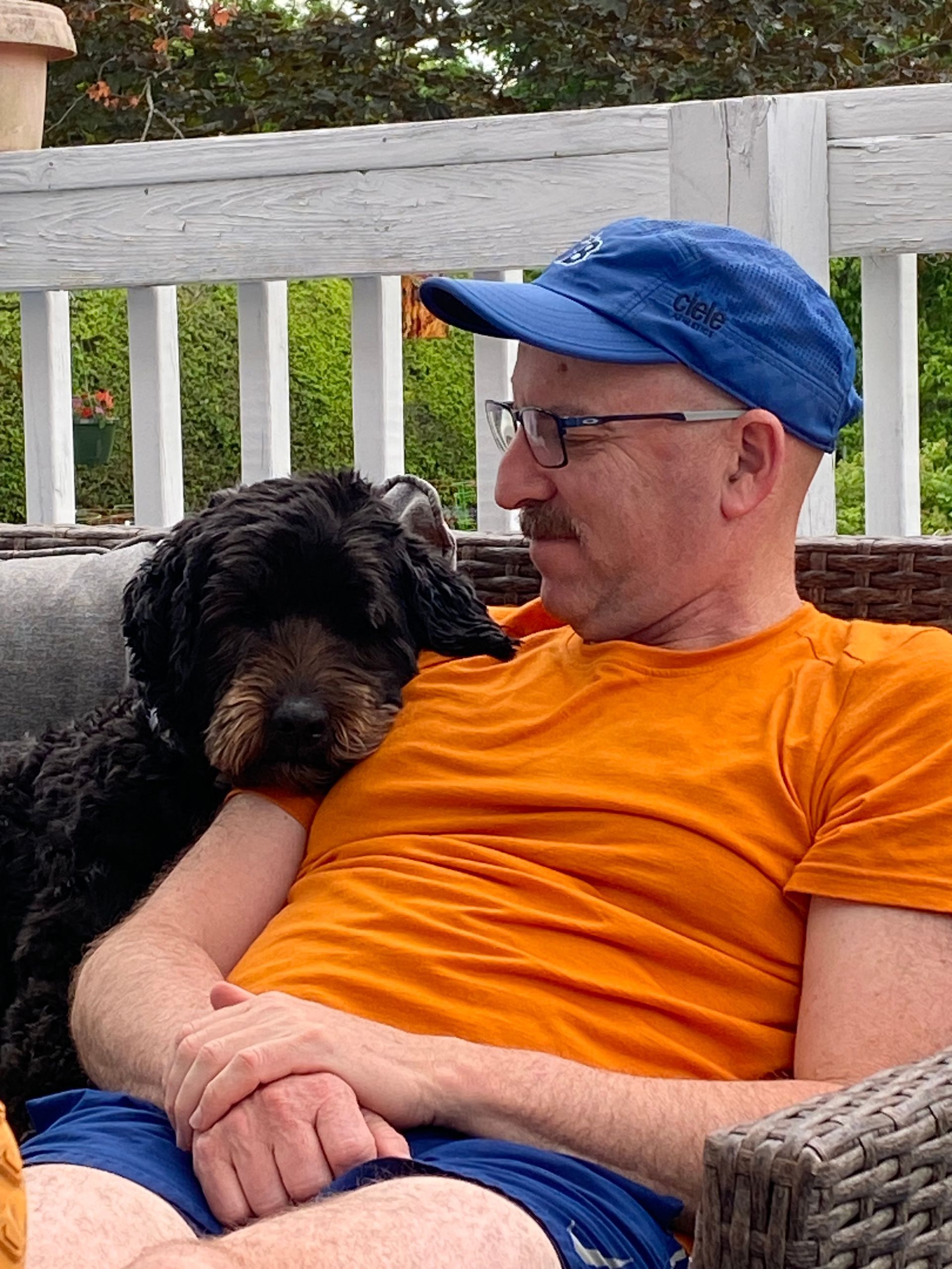 Image shows a man with an orange shirt and blue hat sitting next to a black dog leaning on his shoulder