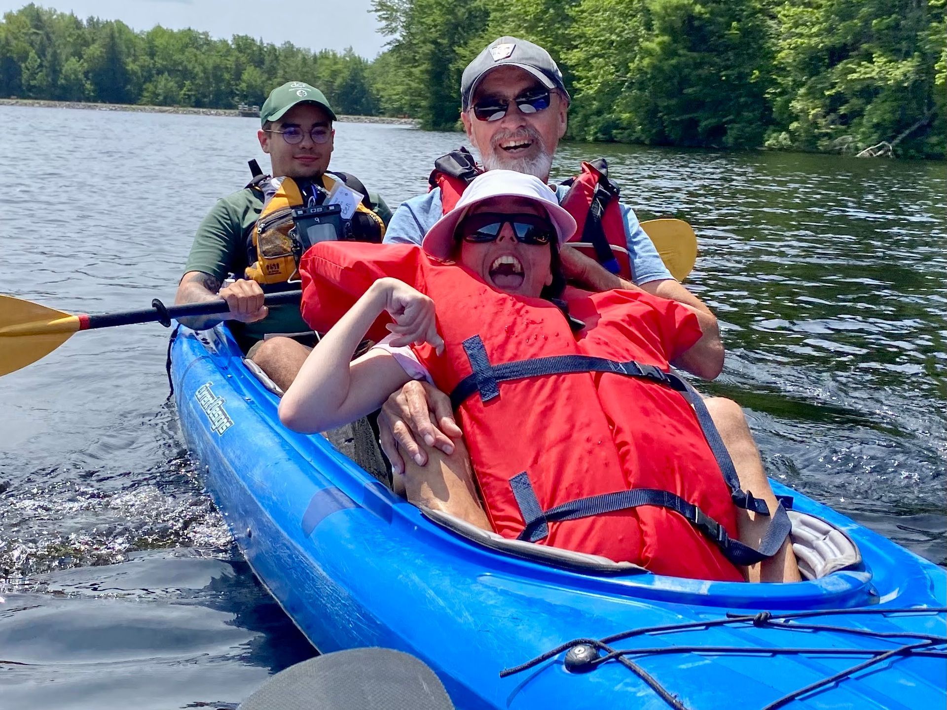 A woman in a red life lacket leans against her father in the front of a blue kayak.  Both have huge smiles.  Behind them, a person is holding a kayak paddle, ready to paddle their kayak.