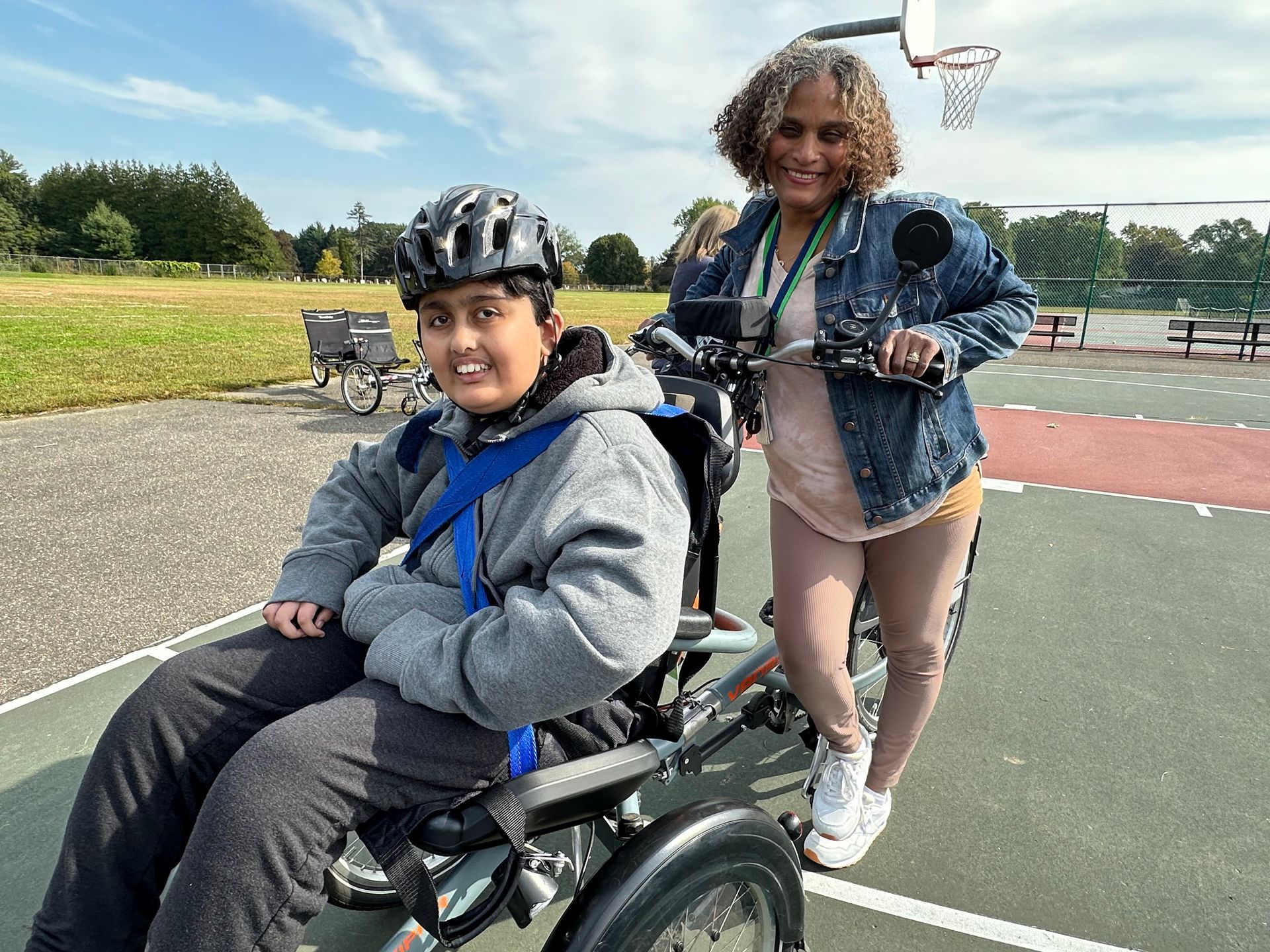 Image shows a teenager using a wheelchair tandem, and an adult behind him, both are smiling at the camera