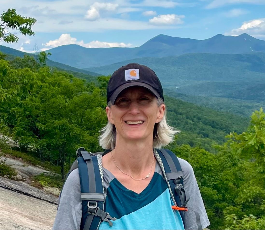 Nicole Rochefort, wearing a black hat and a backpack, smiling at the camera, with dramatic mountains in the background