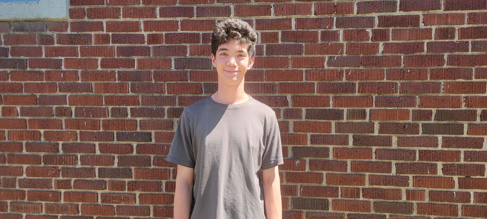 Image shows a young man smiling, posed in front of a brick wall