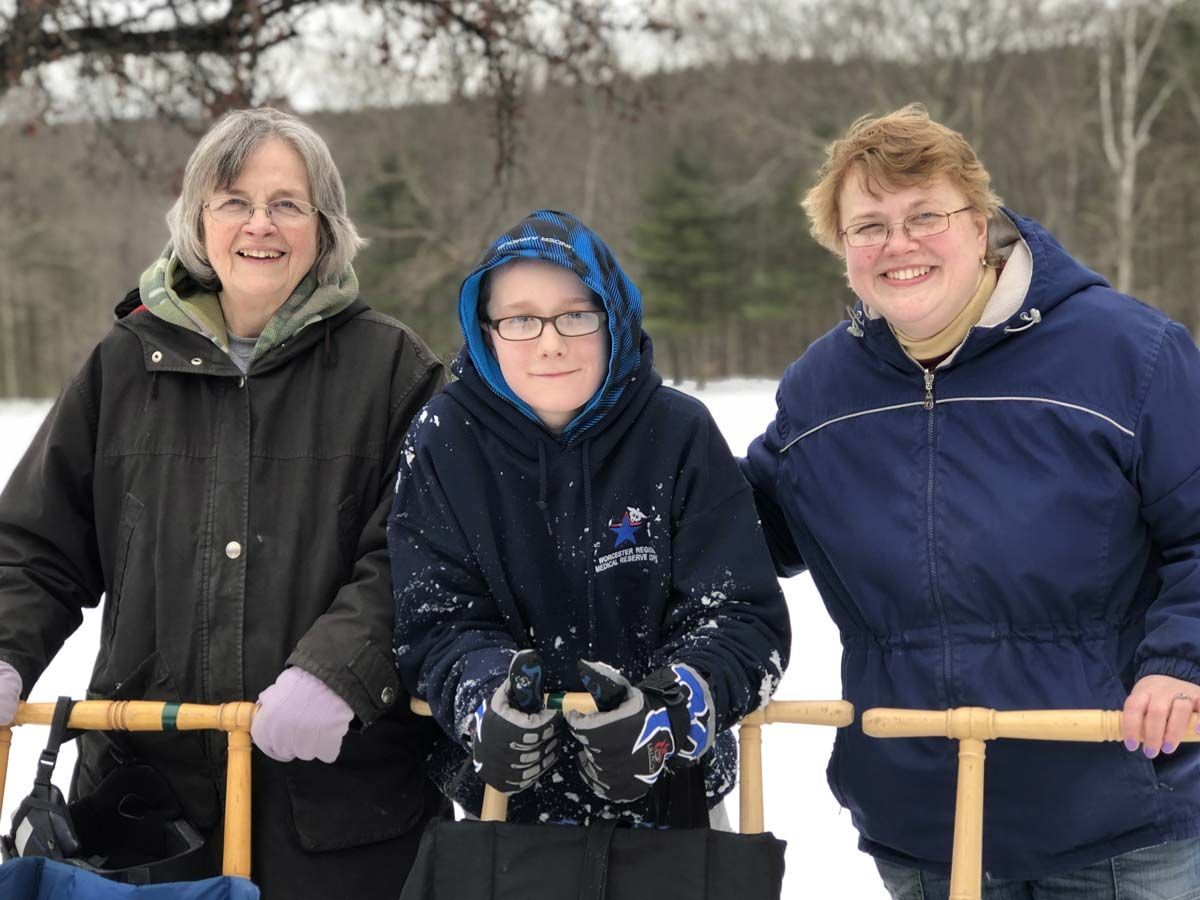 Three people stand in coats in gloves outside, behind kick sleds, smiling at the camera.