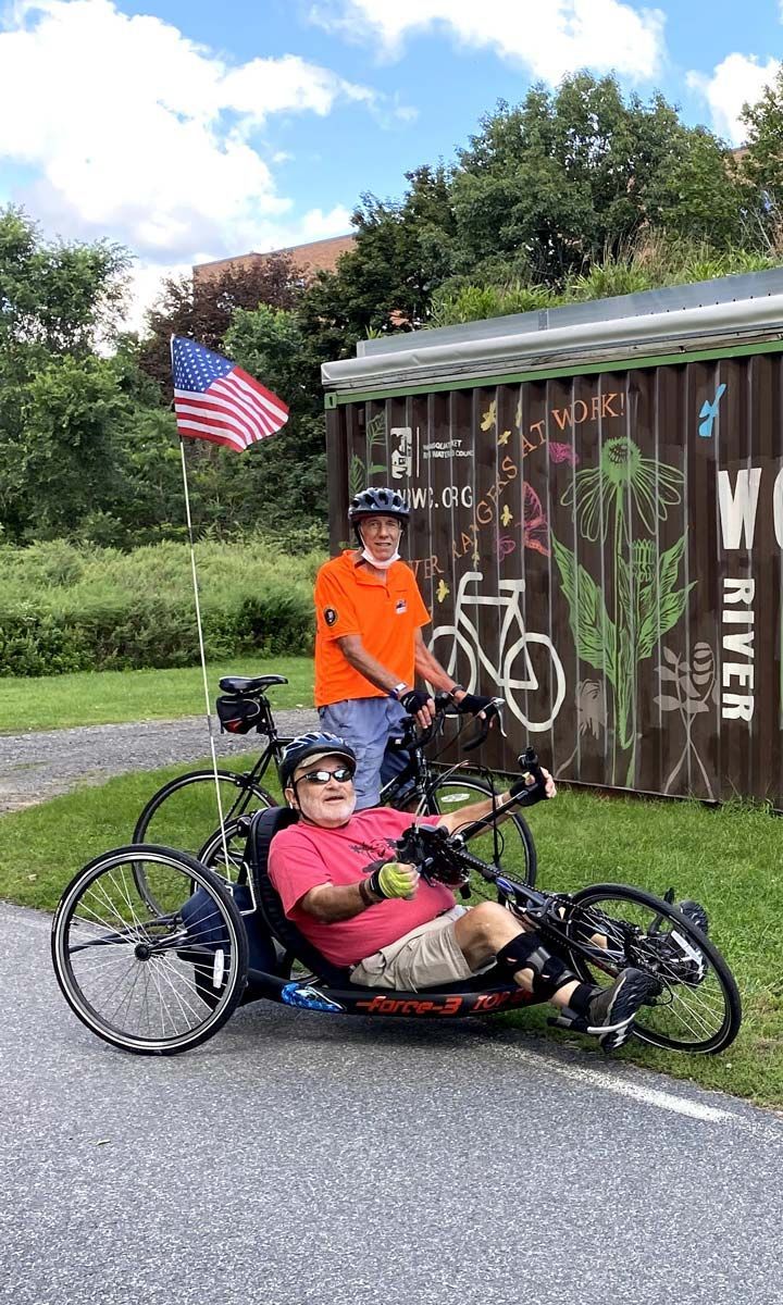 Two gentleman ride on a bicycle and tricycle on the Woonasquatucket River Greenway rail trail.