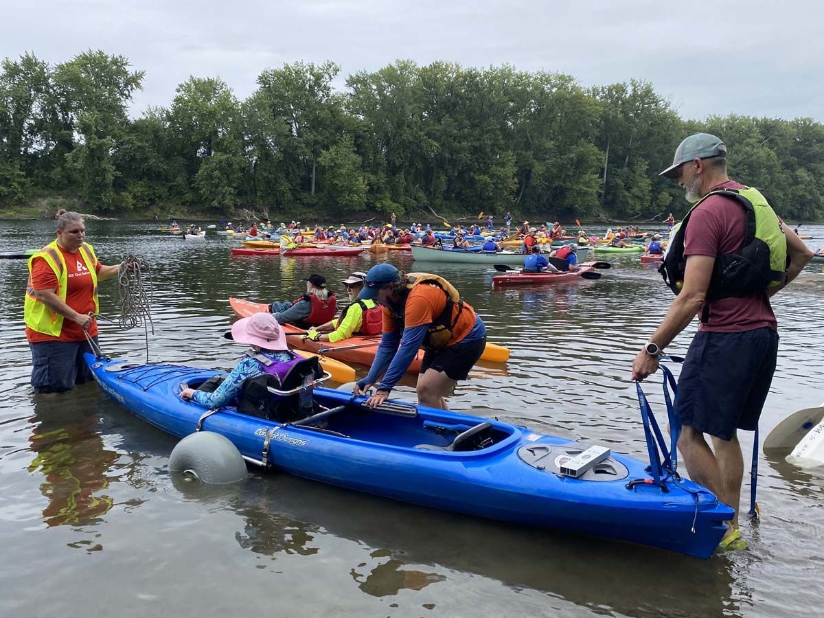 Image shows a woman being helped into a blue tandem kayak while a large flotilla waits behind her
