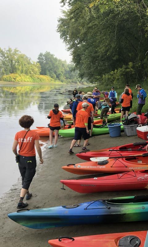 People in t-shirts and shorts standing on a river bank with a row of colorful kayaks.
