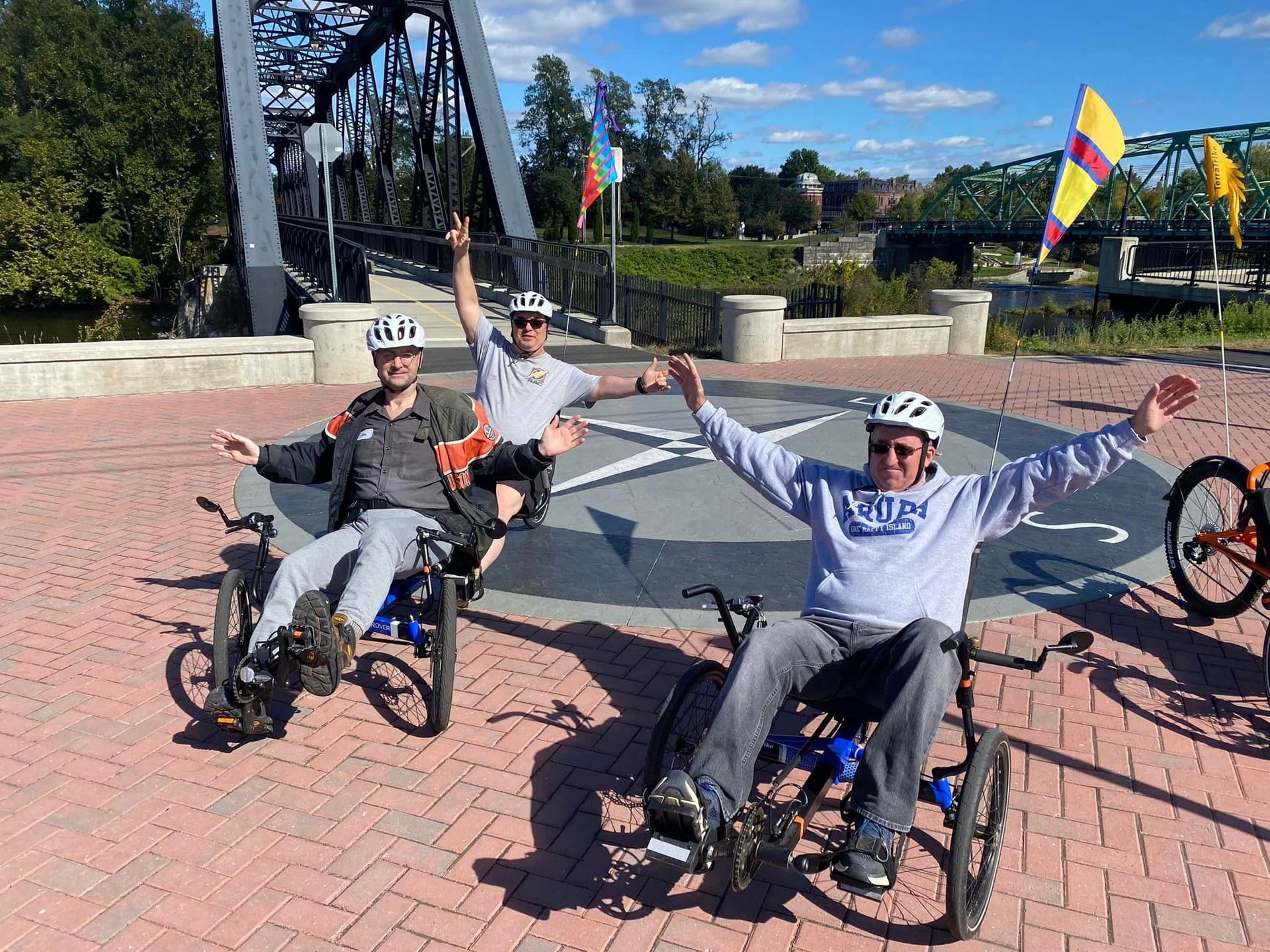 Image shows 4 people on recumbent trikes smiling and waving at the camera in front of a cycling trail bridge