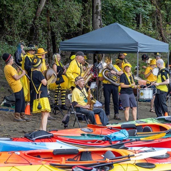 An eclectic band dressed in yellow and black play many instruments on the bank of a river.