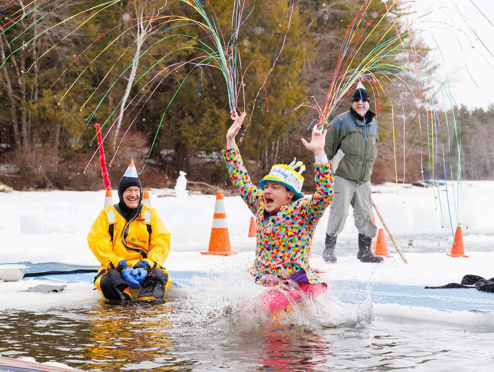 Briggs Doxxzen, wearing a polka dot shirt, a birthday cake hat, and shooting streamers from his hands, plunges into ice cold water while a firefighter laughs in the background