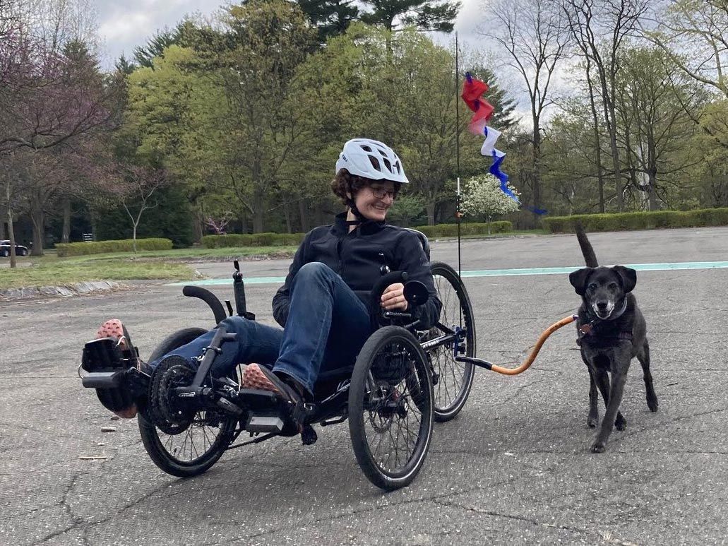 A smiling woman in a helmet rides a recumbent trike accompanied by a black lab.