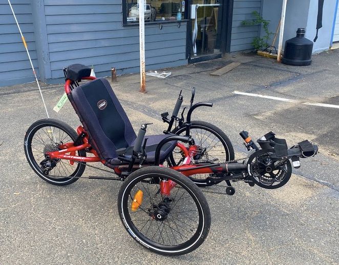 A red recumbent tricycle with reclining seat and vertical handles is parked in front of a building.